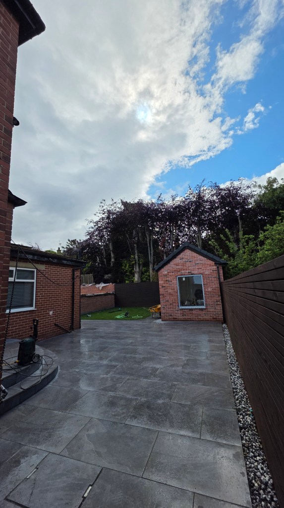 A backyard with a paved patio area, a brick shed with a window, and a green lawn in the background, under a cloudy sky.